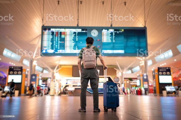 Asian male traveler with wheeled luggage checking for flight schedule on arrival and departure board at Kuala Lumpur International Airport