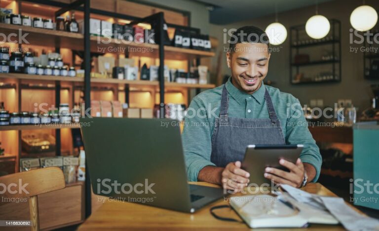 Smiling young male deli owner working a tablet and a laptop while sitting at a table in his artisanal shop